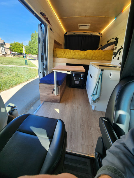Step into this cozy RV rental camper van: bed, dinette, and kitchen. Wood-paneled interior, with an open door looking outside.