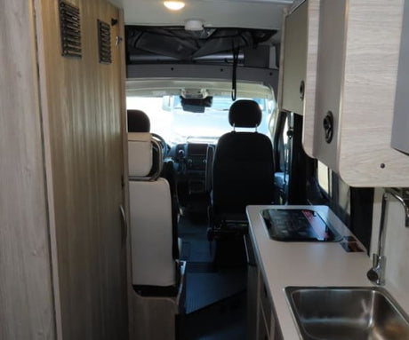 Interior of an rv rental showing a clean kitchenette with sink & cooktop, storage, and the vehicle's front seats.