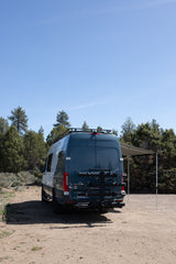 An RV rental, a dark Mercedes Sprinter camper van, parked on a dirt road with its awning extended in a sunny, treed landscape.