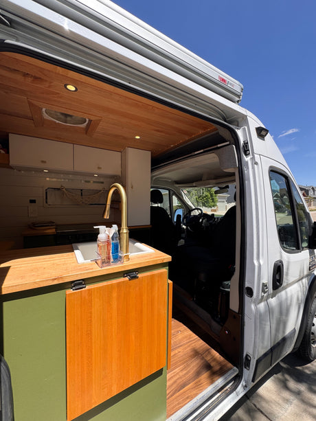 An open-door view inside an RV rental camper van, revealing a custom kitchen with wood counters, a gold faucet, and green cabinets.