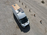 RV rental: Aerial view of a white camper van with a wooden rooftop deck, solar panels, and skylight, parked on a gravel road.