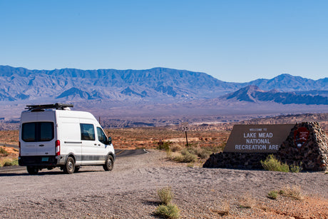 An RV rental van pauses at the entrance to Lake Mead National Recreation Area, with vast desert mountains behind.