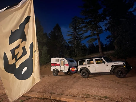 CU flag, white Jeep, & RV rental trailer parked at dusk. Ready for Colorado camping adventure.