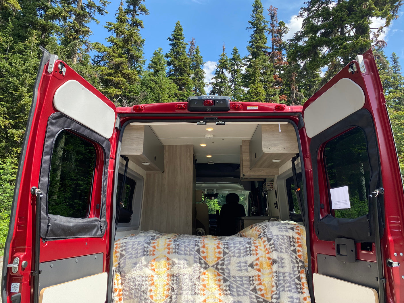 Open rear doors of a red RV rental, revealing a cozy interior bed and a serene forest view.