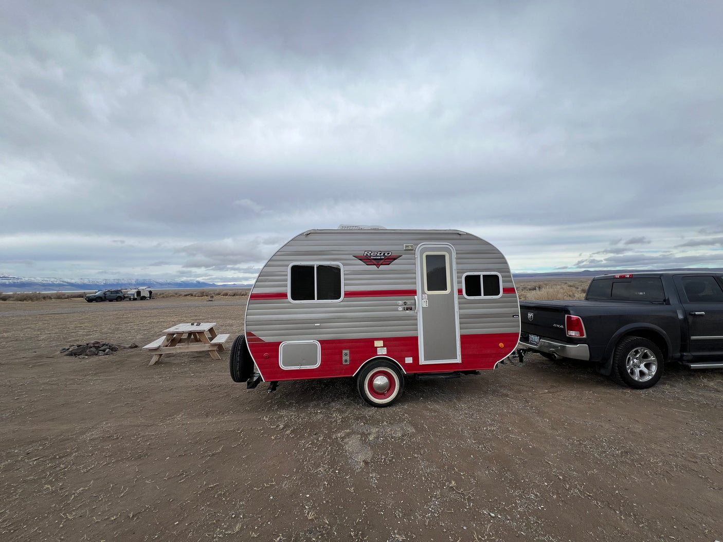 Retro RV rental hitched to a pickup truck, parked by a picnic table in a vast, cloudy landscape with distant mountains.