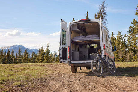 This RV rental camper van is ready for adventure with a mountain bike, parked on a dirt road amidst scenic mountains and forests.