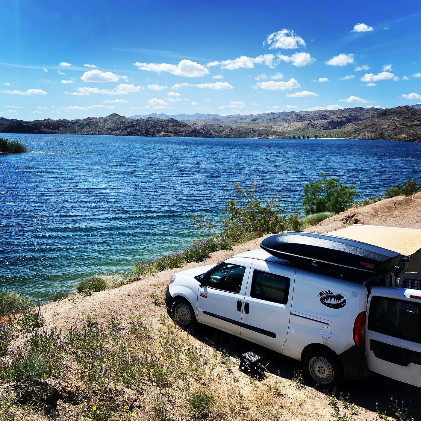 Lakeside white camper van, featuring a roof box & awning, against mountain backdrop. Perfect RV rental for adventure.
