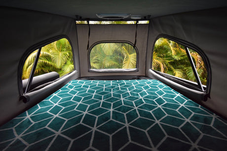 Inside a comfy **RV rental**: cozy bed with teal geometric blanket, offering lush palm tree views from pop-up windows.