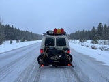 Winter RV rental adventure: White van with motorcycle on snowy road, surrounded by snow-covered pines.