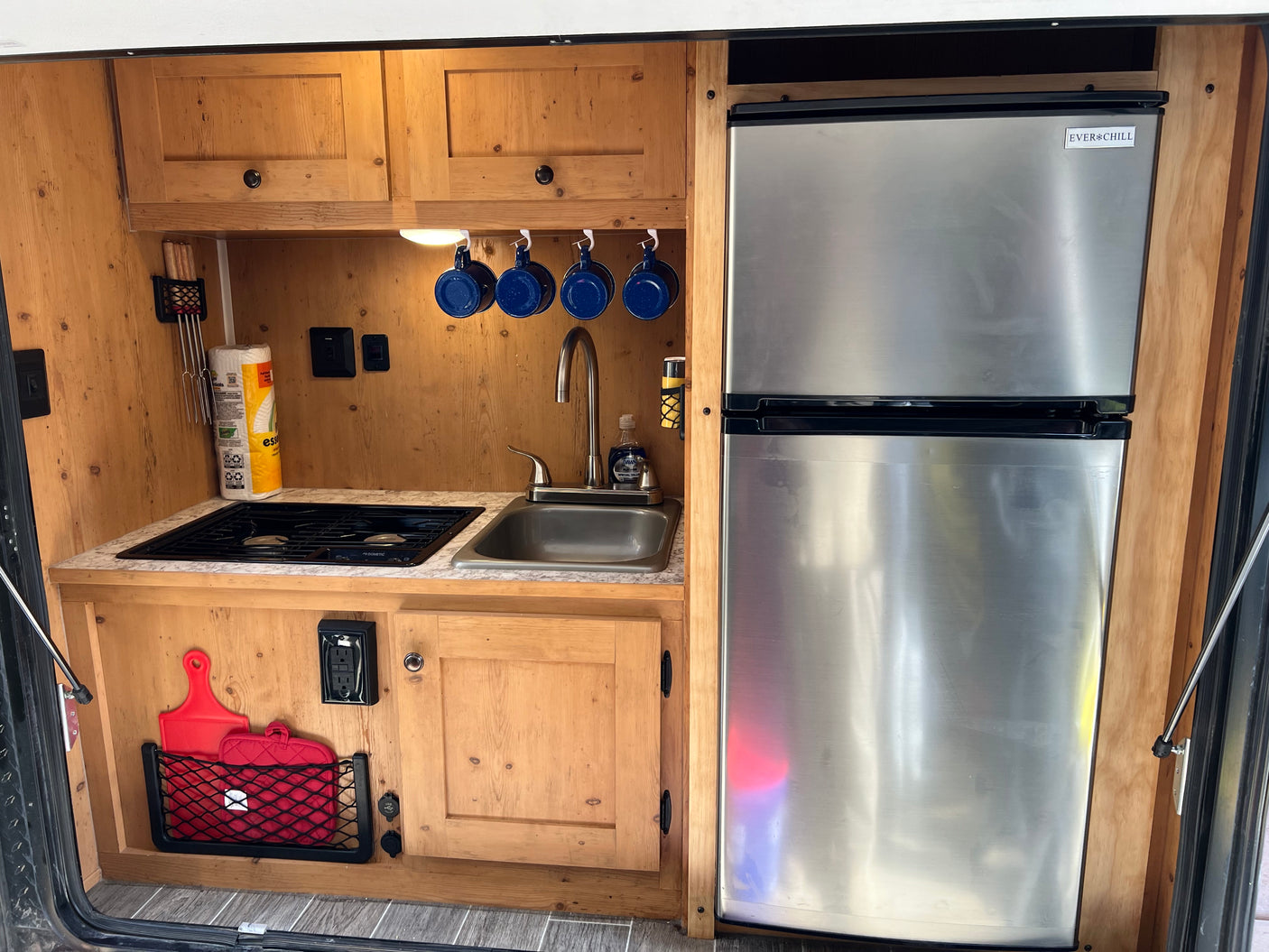 Inside an RV rental, a compact kitchen features wood cabinets, a stainless steel fridge, stove, sink, and essential accessories.