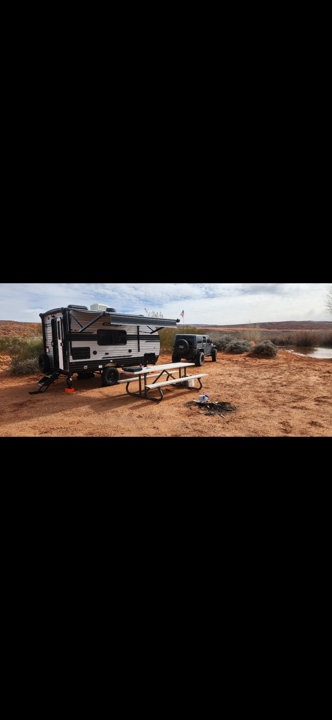An rv rental, Jeep, and picnic table are set up at a red-dirt desert campsite by a small pond.