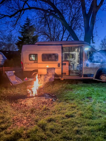 This RV rental adventure features a campervan parked at dusk beside a roaring bonfire and chairs. Cozy night outdoors!