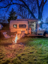 This RV rental adventure features a campervan parked at dusk beside a roaring bonfire and chairs. Cozy night outdoors!