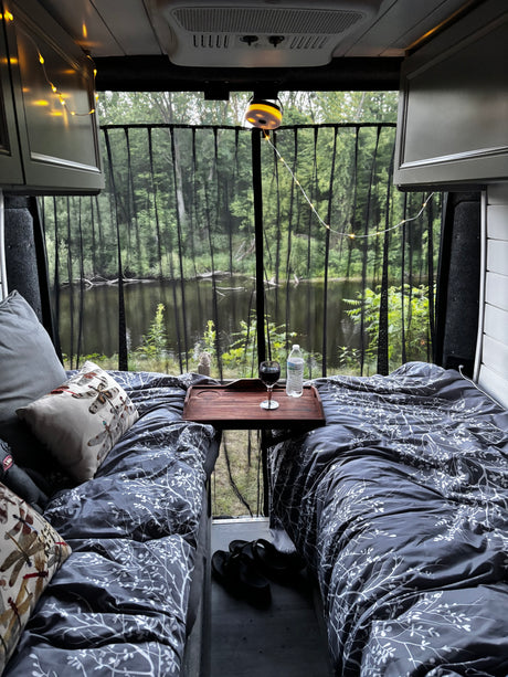 Cozy RV rental bedroom overlooks a forest lake through a mesh screen. Enjoy wine & comfort in nature.