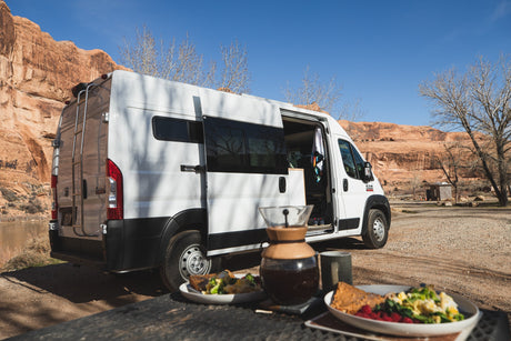 A white RV rental with an open side door is parked by a river, red rock cliffs, and bare trees, with breakfast on a table.