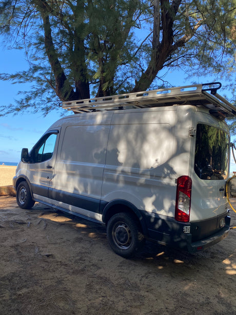 White camper van parked under trees by a sunny beach, ideal for an RV rental getaway.