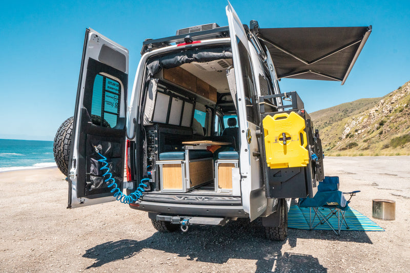 RV rental with open rear doors by the beach, showcasing its adventure-ready interior, gear, and deployed awning. — photo 11