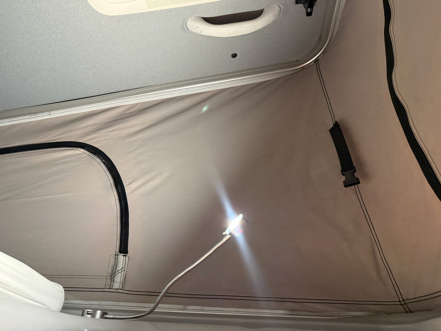 Interior of an RV rental camper van, showing a beige pop-top roof, a bright flexible USB LED light, and the grey vehicle ceiling with a grab handle.