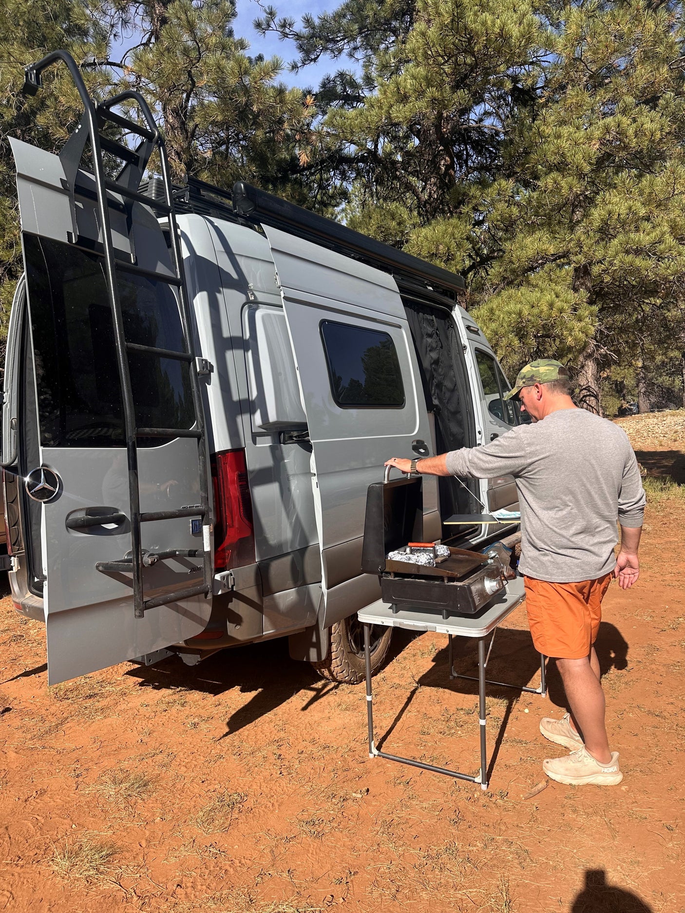 A man cooks outdoors on a griddle by a gray RV rental camper van with open rear doors.