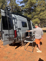 A man cooks outdoors on a griddle by a gray RV rental camper van with open rear doors.
