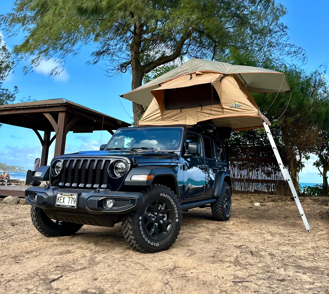 Rooftop tent on a black Jeep Wrangler parked on sand near a beach and gazebo under a tree. Great **rv rental** for adventurous trips. — photo 2