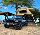 Rooftop tent on a black Jeep Wrangler parked on sand near a beach and gazebo under a tree. Great **rv rental** for adventurous trips.