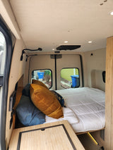 Interior of an RV rental: a cozy bed with pillows, natural wood accents, and bright lighting, visible through the rear windows.