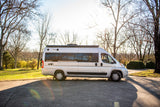 Silver Winnebago Travato camper van rv rental, parked on a sunlit pavement amidst bare trees and green grass.