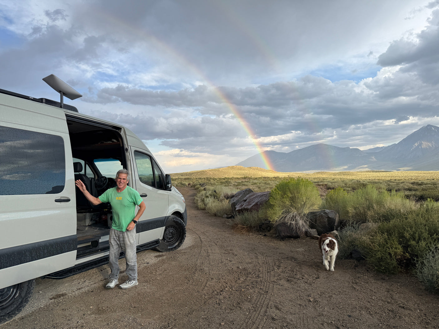 Enjoy your RV rental adventure! Man and dog with a Starlink-equipped van under a rainbow in a scenic landscape.