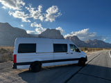 White RV rental camper van parked on a desert road, with large mountains and a cloudy blue sky.