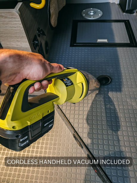 Inside an RV rental, a hand uses a Ryobi cordless handheld vacuum on the gray textured floor. Cleaning convenience included for trips.