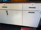RV rental storage: white cabinets, drawers with black handles and travel latches, dark wooden counter, colorful rug on floor.