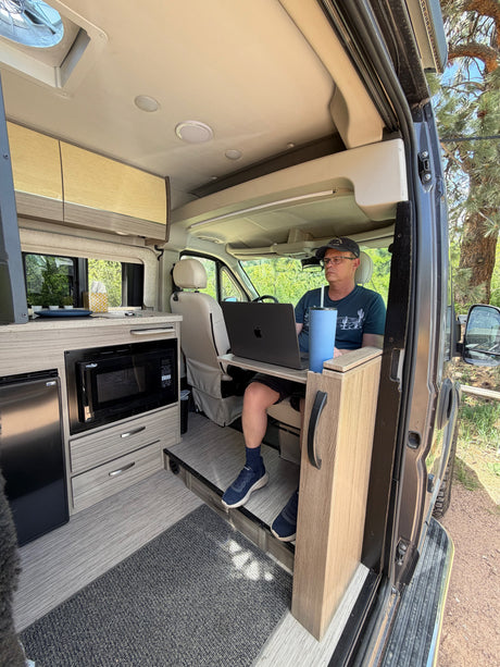 Man in glasses working on laptop inside a modern rv rental van. View of kitchen area with microwave, fridge, and outdoor trees.