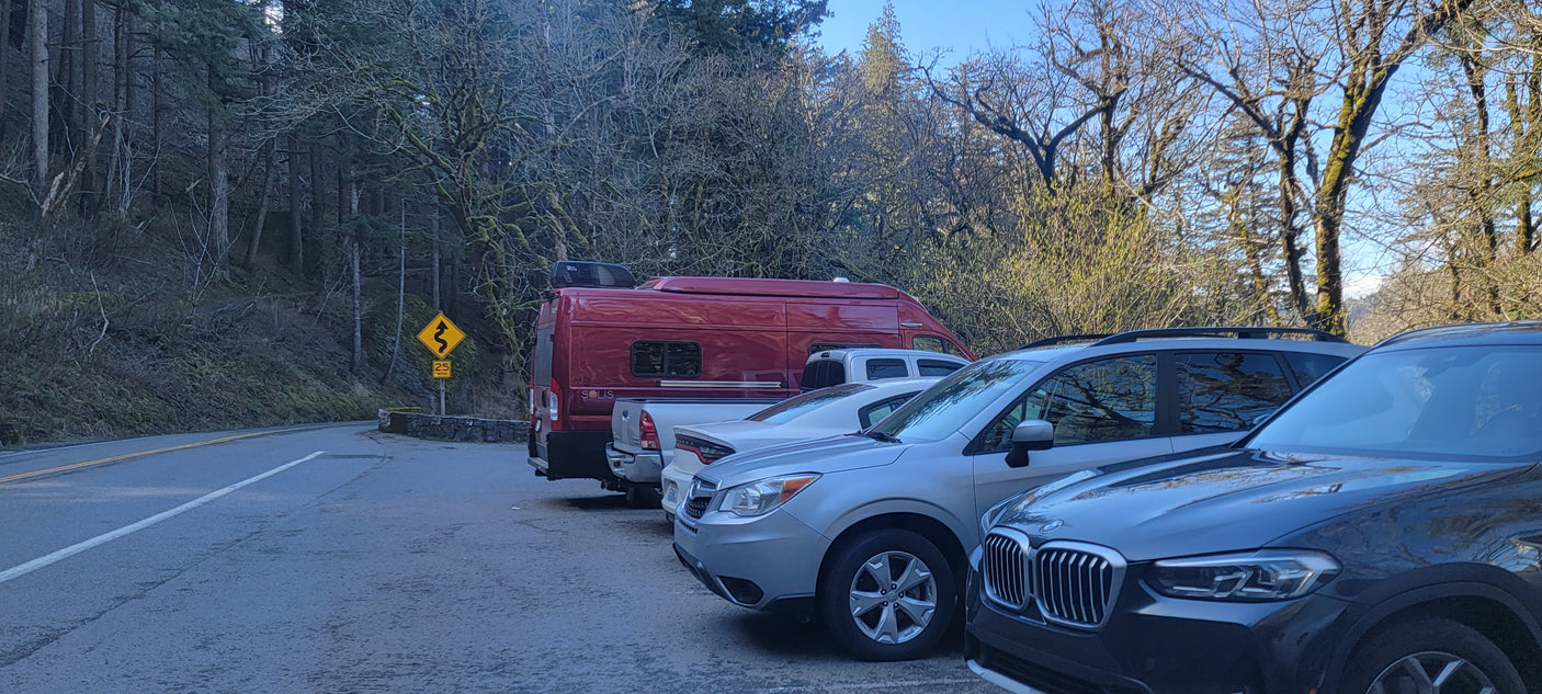 Red RV rental and cars parked in a forest parking lot beside a winding road with a 25 mph sign.