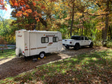 An RV rental and white Jeep Gladiator are parked at a serene autumn campsite, surrounded by trees with vibrant fall foliage.