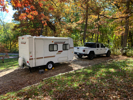 An RV rental and white Jeep Gladiator are parked at a serene autumn campsite, surrounded by trees with vibrant fall foliage.