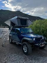 This blue Jeep with a deployed rooftop tent by a lake offers an amazing RV rental experience amidst scenic mountains.