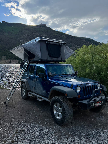 This blue Jeep with a deployed rooftop tent by a lake offers an amazing RV rental experience amidst scenic mountains.