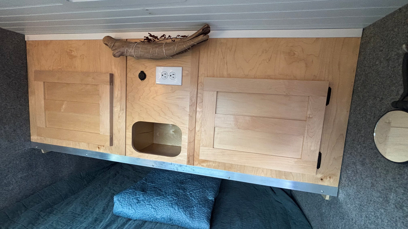 RV rental interior: Light wood overhead cabinets, cubby, and outlets above a dark quilted bed. Gray walls, slatted ceiling.