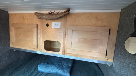 RV rental interior: Light wood overhead cabinets, cubby, and outlets above a dark quilted bed. Gray walls, slatted ceiling.
