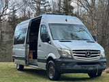 A silver Mercedes-Benz Sprinter camper van, ideal for rv rental, parked on grass with side door open, showing its interior.