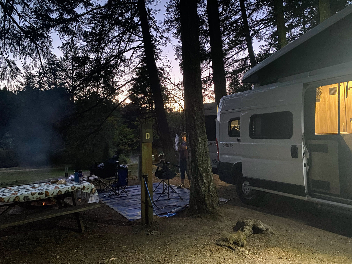 Enjoying a serene evening at a forest campsite with a Winnebago Solis RV rental, campfire glowing, and person busy.