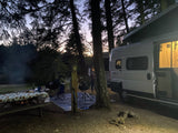 Enjoying a serene evening at a forest campsite with a Winnebago Solis RV rental, campfire glowing, and person busy.