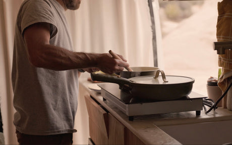 Man cooks in a compact rv rental kitchen, stirring a pot on an induction cooktop with a scenic outdoor view.