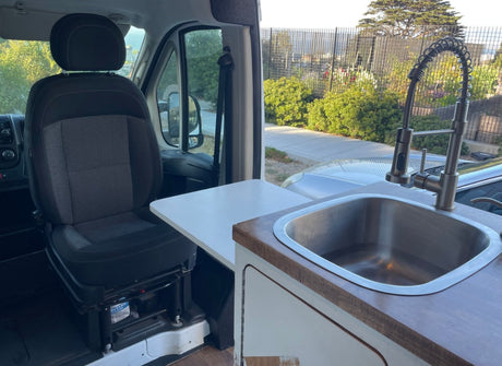 RV rental interior: passenger seat, small table, and a kitchen sink with modern faucet. Street view visible outside.