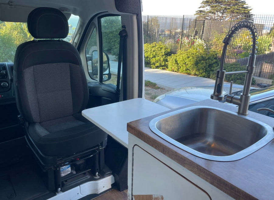 RV rental interior: passenger seat, small table, and a kitchen sink with modern faucet. Street view visible outside.