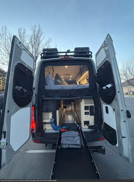 RV rental: Back of a camper van with open doors, showing a cozy bed, interior lighting, and a pull-out ramp for gear.