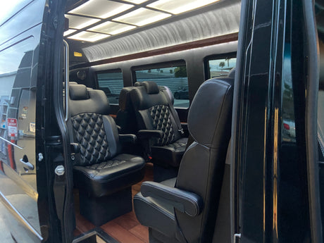 Interior of a luxury RV rental van with black quilted leather captain's chairs, wood floor, and overhead lighting.