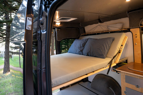 View inside an RV rental camper van: A comfortable bed with grey pillows and wooden cabinetry, seen through the open side door in nature.