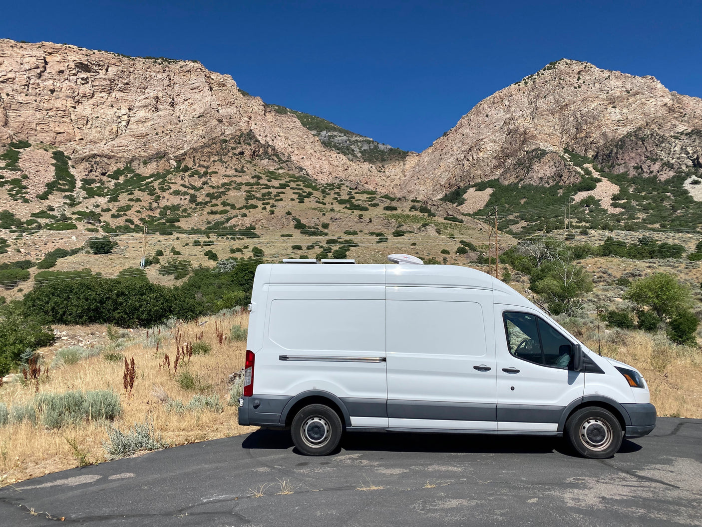 White Ford Transit rv rental camper van, equipped with an AC unit, parked on asphalt before rocky mountains and clear sky.